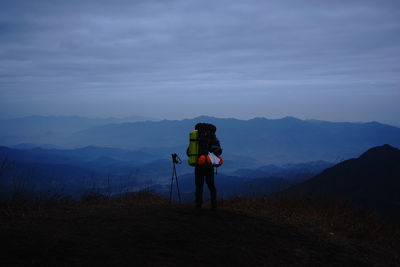 Rear view of person with backpack standing on mountain against sky at dusk