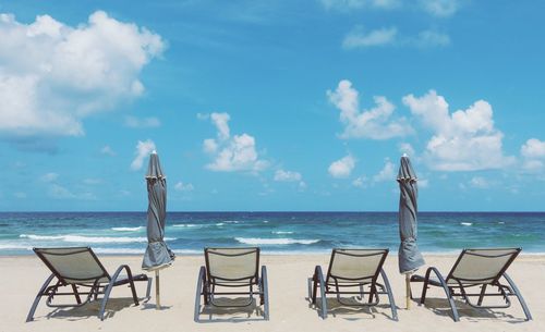 Chairs and closed parasols at beach against sky