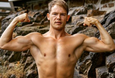 Portrait of shirtless man flexing muscles while standing against rock formation