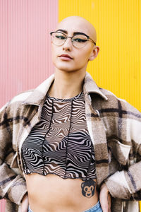 Young woman with shaved head wearing eyeglasses in front of wall