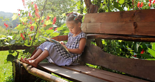 Cute 6-7 years old school girl reading an abc book on the bench in the summer garden in village