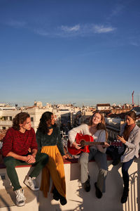 Young woman playing acoustic guitar to cheerful young multiracial friends while having party on sunny rooftop