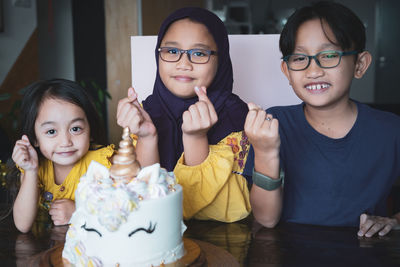 Portrait of smiling siblings with cake on table gesturing heart shape