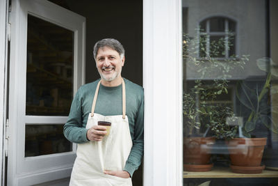 Portrait of confident mature male owner having coffee at doorway of art studio