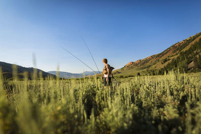 Teenage boy holding fishing rods while standing on field against clear sky