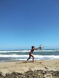 Full length of woman playing with dog on shore at beach against clear blue sky