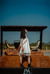 Full length of young woman standing on rusty vehicle against clear sky
