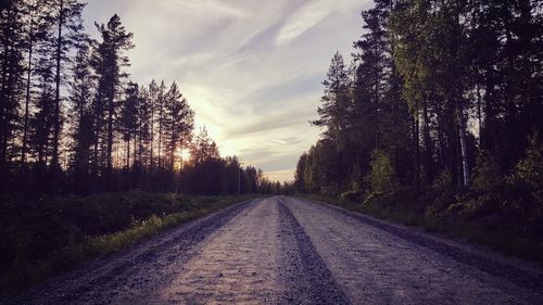 Dirt road amidst trees against sky during sunset