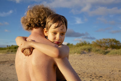 Cute boy embracing shirtless father at beach against sky