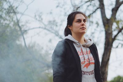 Woman looking away while standing against tree during foggy weather