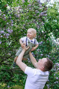 Young father is holding his little son on hands on background of blossom lilac flowers trees.