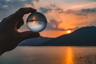 Person holding crystal ball against sea during sunset