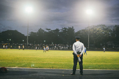 Rear view of security guard looking at players playing soccer on field at night