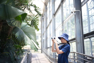 Side view of man holding camera while standing by tree