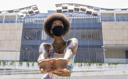 Low angle of african american female athlete in protective mask warming up arms before workout in city during coronavirus pandemic