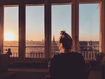 Rear view of woman looking through window at sunset