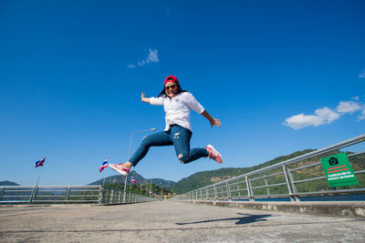 Full length of woman jumping on bridge against blue sky