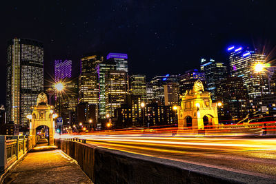 Light trails on street amidst illuminated buildings against sky at night