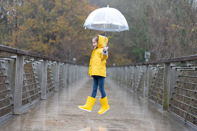 Full body side view of cheerful little girl in bright yellow raincoat and rubber boots with umbrella in hand jumping on wet bridge in rainy autumn weather in park