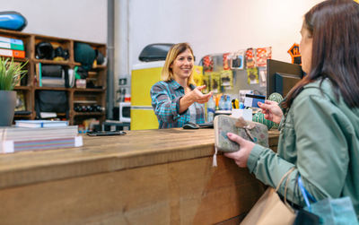 Female customer giving credit card to smiling woman shop assistant to pay purchase on local store