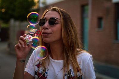 Young woman wearing sunglasses blowing bubbles while standing on street