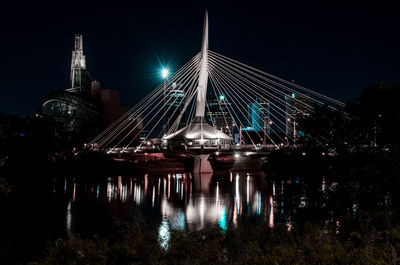 Illuminated bridge over river by buildings against sky at night