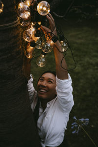 Smiling female hanging lighting equipment on tree trunk in yard during dinner party