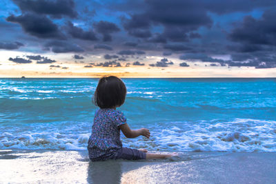 Boy sitting on beach against sky during sunset