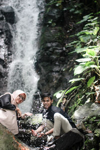 Man and woman splashing water against waterfall in forest
