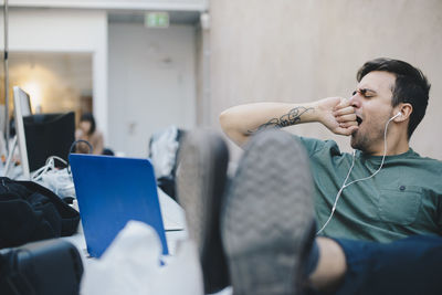 Tired computer programmer yawning while sitting with feet up at desk in office