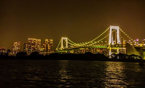 Illuminated suspension bridge over river against sky at night