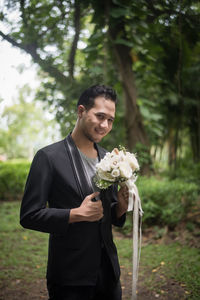 Portrait of groom holding flower bouquet while standing on grass in forest