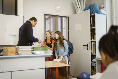 Mature male teacher giving documents to students standing in classroom