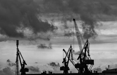 Silhouette cranes at harbor against cloudy sky