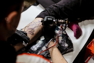 From above cropped unrecognizable man with using tattoo machine to make tattoo on leg of crop customer during work in salon