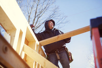 Low angle view of worker constructing wooden house against blue sky