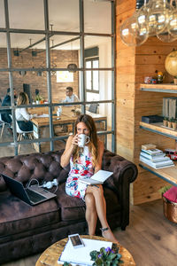 Young woman drinking coffee while rading book on sofa against people