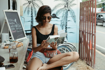 Resting woman in casual wear and trendy sunglasses reading book during refreshment in outdoors bar