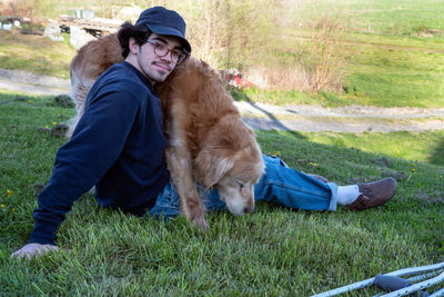 A young man on a grassy hill  cuddled by loving golden retriever dog with crutches in foreground 