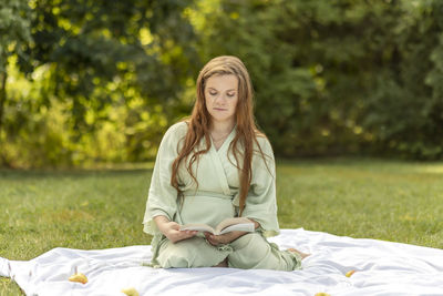 Young pretty pregnant girl holding, reading a book,resting in park,sitting on cover,blanket in park
