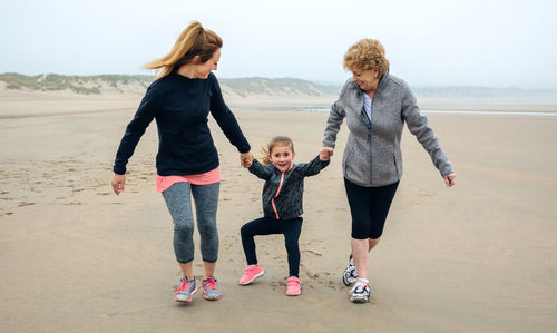 Playful woman walking with mother and daughter at beach against sky