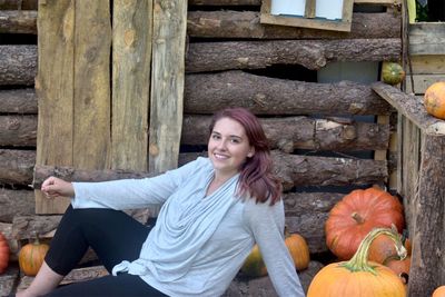 Portrait of smiling young woman sitting by pumpkins against log cabin