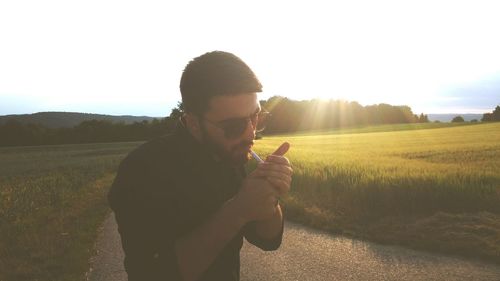 Man igniting cigarette while standing on grass against sky