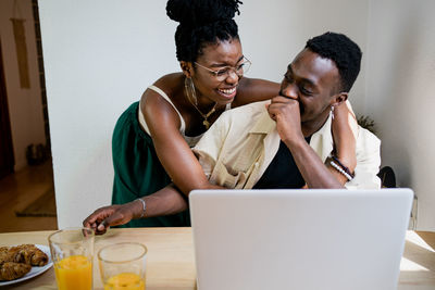 Cheerful african american female in crop top hugging positive husband working at laptop remotely in bright cozy studio apartment