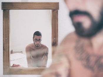 Portrait of shirtless man reflecting in mirror with friend in foreground