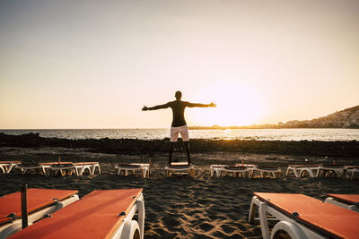 Rear view of silhouette young man with arms outstretched standing on deck chair at beach against clear sky during sunset