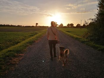 Rear view of woman walking with dog on road against sky during sunset