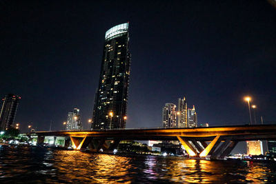 Illuminated bridge over river by buildings against sky at night