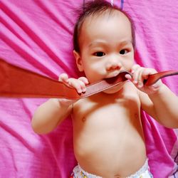 Directly above portrait of cute baby boy biting belt while lying on pink bed