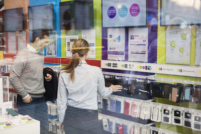 Saleswoman helping customer in buying phone cover seen through glass